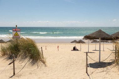Portekiz 'in Costa da Caparica şehrinin altın kumlarına gölge düşüren saman plajı şemsiyeleri. Parlak bir gökyüzü altında sakin bir deniz manzarası, rahatlama ve yaz tatili görüntüsü için mükemmel..