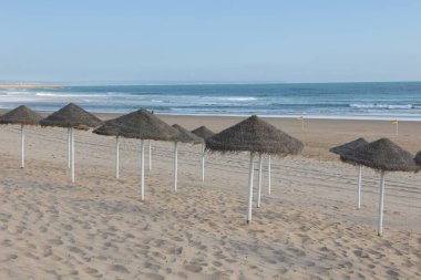 Portekiz 'deki Costa da Caparica plajının manzarası. Altın kum üzerinde saman şemsiyeler, arka planda sakin mavi bir okyanus ve açık bir gökyüzü. Mükemmel huzurlu yaz seyahati.