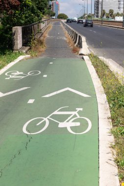 Yeşil asfalt bir yol üzerinde beyaz bisiklet sembolleri ve oklarla işaretlenmiş bisiklet yolu çimen ve bitki örtüsüyle çevrili, Portekiz 'de kentsel bisikletçiliği ve güvenli ulaşımı teşvik ediyor