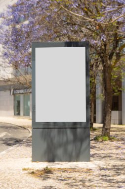 Baharda Lizbon caddesinde boş bir ilan panosu, çiçek açmış jakaranda ağaçlarıyla çevrili. Modern bir şehirde doğal ışık ve mevsimlik cazibe ile reklam için ideal şehir modeli..