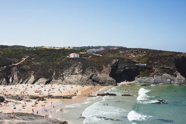 Zambujeira do Mar 'daki kayalık uçurumlarla çevrili manzaralı plajda insanlar güneşin ve okyanusun keyfini çıkarıyorlar. Açık mavi gökyüzü ve turkuaz sular mükemmel bir yaz tatili atmosferi yaratır..