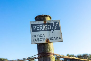Açık mavi gökyüzünün altındaki tahta direkte Perigo Cerca Electrificada yazıyor. Kırsal bölgede paslı ve yıpranmış dokuları olan elektrikli çitlere dikkat edin..