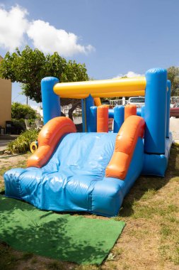 Renkli şişme kaydırak güneşli bir günde açık havada kurulur. Çocuk partileri, bahçe eğlencesi ve yaz eğlencesi için ideal. Mavi, turuncu ve sarı yapılar yumuşak, güvenli malzemelerle.