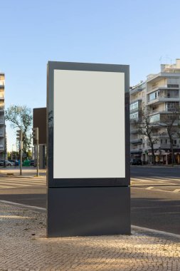 Gündüz vakti bir cadde kavşağındaki kaldırımda boş bir reklam panosu modeli. Boş reklam alanı özel içerik için hazır, kentsel binalar ve arka planda açık gökyüzü.
