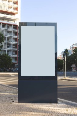 Şehir kaldırımında boş dikey bir ilan panosu duruyor. Etrafı uzun binalar, ağaçlar ve günışığında sakin bir sokak ile çevrili..