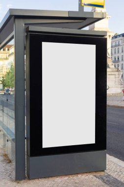 Günümüz otobüs durağında boş bir reklam panosu, güneşli bir öğleden sonra klasik binalar, hafif trafik ve ağaçlarla çevrili bir Avrupa şehrinin kaldırımında duruyor..