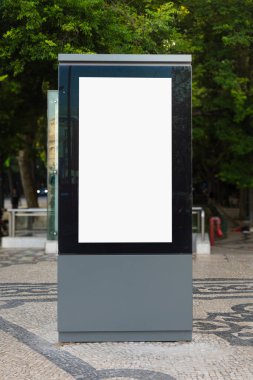 Dikey boş bir reklam panosu, ağaçlar ve şehir binalarıyla çevrili taşlarla kaplı kaldırımda duruyor. Modern bir metropol bölgesinde kentsel reklam için hazır..