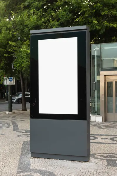 Dikey boş bir reklam panosu, ağaçlar ve şehir binalarıyla çevrili taşlarla kaplı kaldırımda duruyor. Modern bir metropol bölgesinde kentsel reklam için hazır..