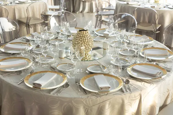 Resmi bir etkinlik ya da düğün için zarif bir ziyafet masası, altın çerçeveli tabaklar, katlanmış peçeteler, kristal cam süsler ve yumuşak doğal ışık altında dekoratif altın bir süs..