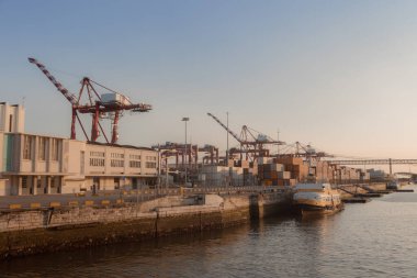 Gün batımında Lizbon Limanı 'nda nakliye konteynırları ve vinçler. Kargo operasyonları ve sakin sularla sanayi sahnesi.