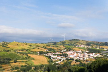 Portekiz 'in Mafra kentindeki kırsal manzarada kayan tepeler, üzüm bağları, rüzgar türbinleri ve beyaz evleri ve kırmızı çatıları olan küçük bir köy yer alıyor. Sürdürülebilir enerji kırsal cazibeyle harmanlanır.