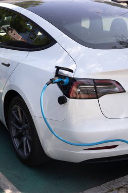 Beyaz elektrikli bir arabanın arka tarafında, muhtemelen bir Tesla Model 3, parlak mavi kabloyla bir EV şarj istasyonuna bağlı. Araç belirlenmiş bir şarj yerine park edilmiş.