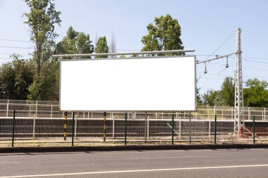  Arka planda tren yolu ya da tramvay pisti olan bir yol kenarına yerleştirilmiş büyük boş bir reklam panosu. Metal bir çit ve elektrik kablolarıyla çevrili bu temiz, boş reklam alanı yüksek görüş imkanı sunuyor.