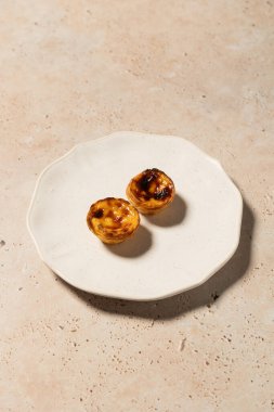 Pastis de nata olarak bilinen iki geleneksel Portekiz kremalı turtasının üst görüntüsü bej taşı bir yüzey üzerinde beyaz bir seramik tabağa yerleştirilmiştir..
