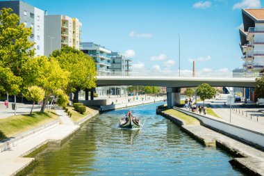 Portekiz 'in Aveiro kentindeki bir kanalın manzarası, güneşli bir günde çağdaş binalar ve yeşil kentsel alanlarla çevrili modern bir köprünün altında geleneksel bir moliceiro teknesi yüzüyor..