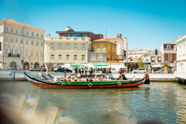 Turistler, Aveiro, Portekiz kanalları boyunca, 2025 tarihli renkli kurdelelerle kaplı köprünün yakınında tekne turnesinin keyfini çıkarıyorlar.