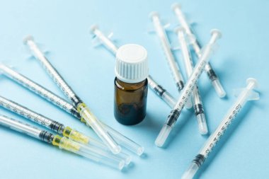 Açık mavi arka planda küçük bir kahverengi ilaç şişesinin etrafına yerleştirilmiş birden fazla tek kullanımlık şırınga tıbbi tedavi, aşı ya da ilaç tedavisini temsil ediyor..