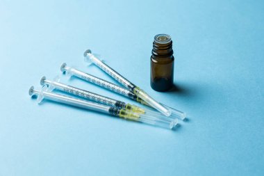 Tıbbi bir şırınganın yakın plan görüntüsü, kapağı çıkarılmış kahverengi bir cam şişe ve açık mavi bir arkaplan üzerine yerleştirilmiş plastik bir iğne kapağı. Kurulum, sağlık hizmetlerini temsil eden bir aşı veya tıbbi enjeksiyon senaryosu öneriyor