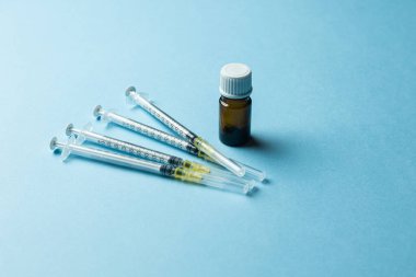 Tıbbi bir şırınganın yakın plan görüntüsü, kapağı çıkarılmış kahverengi bir cam şişe ve açık mavi bir arkaplan üzerine yerleştirilmiş plastik bir iğne kapağı. Kurulum, sağlık hizmetlerini temsil eden bir aşı veya tıbbi enjeksiyon senaryosu öneriyor