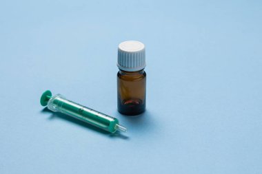 Tıbbi bir şırınganın yakın plan görüntüsü, kapağı çıkarılmış kahverengi bir cam şişe ve açık mavi bir arkaplan üzerine yerleştirilmiş plastik bir iğne kapağı. Kurulum, sağlık hizmetlerini temsil eden bir aşı veya tıbbi enjeksiyon senaryosu öneriyor