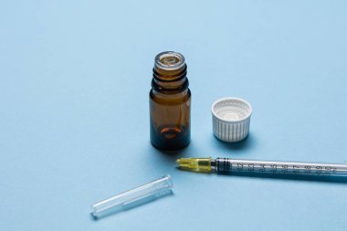 Tıbbi bir şırınganın yakın plan görüntüsü, kapağı çıkarılmış kahverengi bir cam şişe ve açık mavi bir arkaplan üzerine yerleştirilmiş plastik bir iğne kapağı. Kurulum, sağlık hizmetlerini temsil eden bir aşı veya tıbbi enjeksiyon senaryosu öneriyor