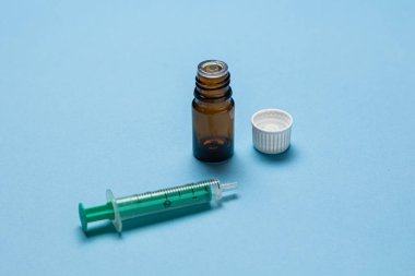Tıbbi bir şırınganın yakın plan görüntüsü, kapağı çıkarılmış kahverengi bir cam şişe ve açık mavi bir arkaplan üzerine yerleştirilmiş plastik bir iğne kapağı. Kurulum, sağlık hizmetlerini temsil eden bir aşı veya tıbbi enjeksiyon senaryosu öneriyor