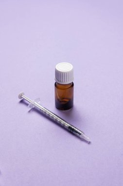 Tıbbi bir şırınga ve açık mor arka planda beyaz kapaklı küçük bir kehribar cam şişe yakın plan, sağlık, aşı veya laboratuvar kullanımını temsil eder.