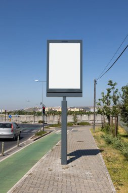Yol kenarındaki kaldırımda dikey boş bir ilan tahtası, yeşil bisiklet yolu ve açık mavi gökyüzünün altında kentsel bir arka plan.