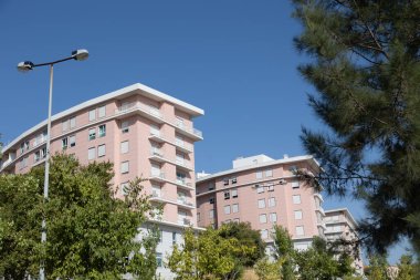 Yeşil ağaçlarla çevrili, balkonları ve beyaz detayları olan modern pembe apartmanlar.