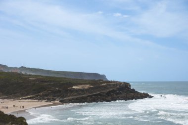 Coastal landscape with sandy beach, cliffs, and ocean waves under a blue sky.