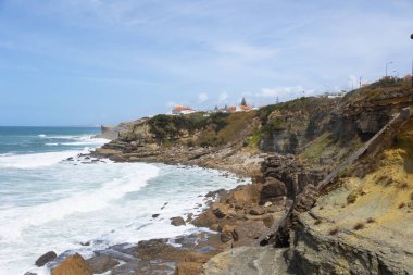 Coastal landscape with sandy beach, cliffs, and ocean waves under a blue sky.
