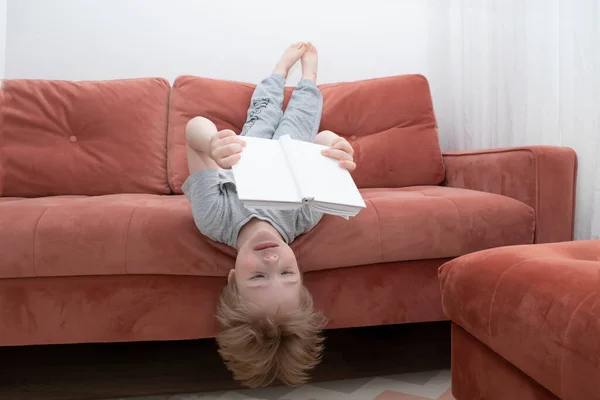 The boy is lying upside down on the sofa and reading a book in a white cover. How to teach a child to read and love books. Educational and entertaining childrens concept