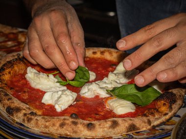 Taze pişmiş bir pizzayı fesleğen ve mozzarella ile ustalıkla süsleyen eller.