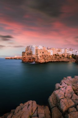 Polignano a Mare, İtalya - Ağustos 2021: Polignano a Mare 'nin günbatımında uzun süre sahnelenmesi, denizin karşısındaki panoramik açıdan görülüyor. Sıcak akşam ışığı ve pürüzsüz suyu olan kayalıklardaki tarihi köy