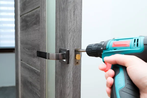 stock image A man repairing a doorknob using a screwdriver at home. Carpenter at lock installation with electric drill into interior wood door