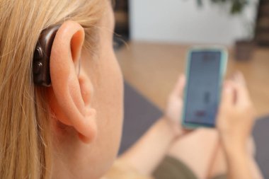 Genç İşitme engelli kadın kapalı alanda akıllı telefon kullanıyor. Kulaklığa işitme cihazı takan ve evde akıllı telefon kullanırken düğmeye basan sarışın kadını kapat..