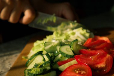 Sebze salatası pişiriyorum. Sebze doğrama işlemi. Kadın elleri mutfakta salatalık ve domates doğruyor. Doğal güneş ışığı, koyu arkaplan