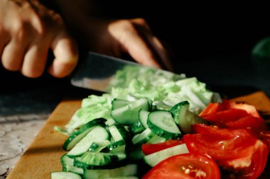 Sebze salatası pişiriyorum. Sebze doğrama işlemi. Kadın elleri mutfakta salatalık ve domates doğruyor. Doğal güneş ışığı, koyu arkaplan
