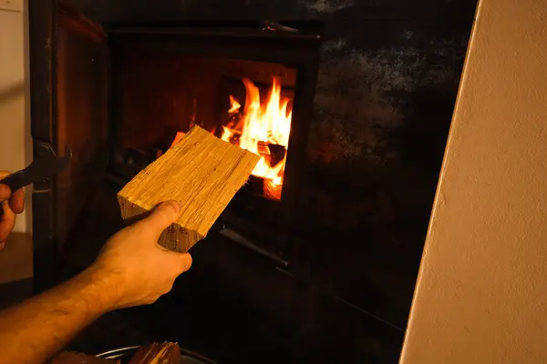 Man putting wood into a fire place. Hand with a log near the open fireplace door. Renewable energy sources.
