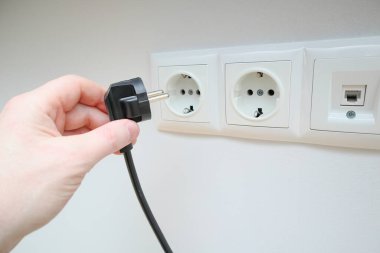 Bir adamın eli beyaz duvardaki prizlerin yanında bir elektrik prizi tutuyor. İnsan eli elektrik prizlerine bir fiş takar. El prizi takar.