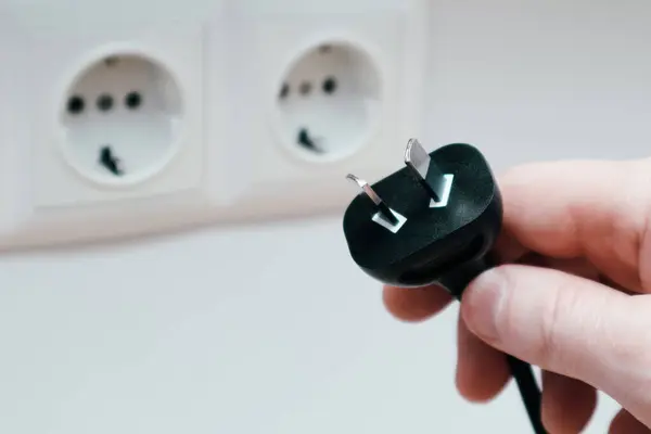 A man's hand holds an electrical plug next to the sockets on the white wall. different standards of electrical plugs and sockets