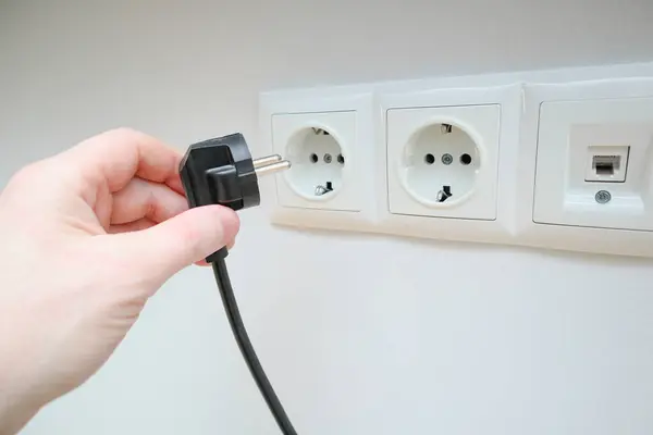 A man\'s hand holds an electrical plug next to the sockets on the white wall. Human hand inserts a plug into an electrical outlets. Hand puts plug in the socket