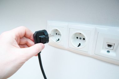 Bir adamın eli beyaz duvardaki prizlerin yanında bir elektrik prizi tutuyor. Eller elektrik prizini tutar.