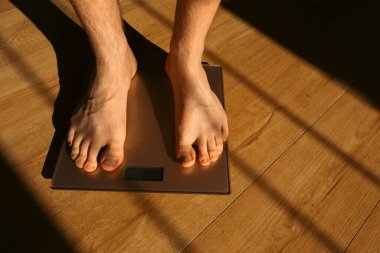 Erkek tartıda ağırlığını kontrol ediyor. Banyo üstüne ağırlık kontrolü için ayaklar. Evde ağırlık tartısı üzerinde duran bir adam. Güneş gölgeleri.