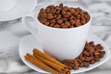 Beyaz fincanda kavrulmuş kahve çekirdekleri ve kokulu tarçın çubukları.