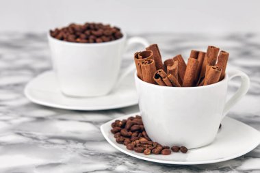 Beyaz fincanda kavrulmuş kahve çekirdekleri ve kokulu tarçın çubukları.