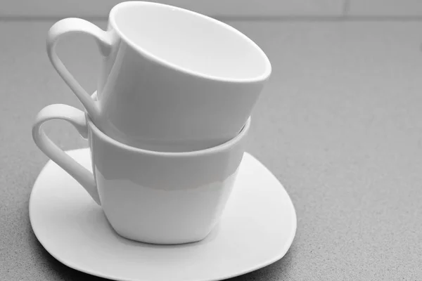 two white coffee mugs standing one on top of the other on a white saucer in black and white