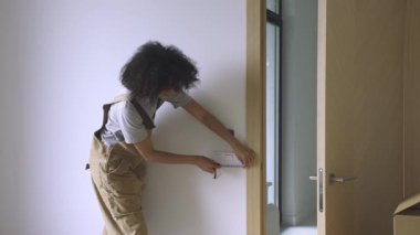 Bir ölçüm bandı kullanan genç bir kadın, güneşli, yenilenmiş bir iç mekandaki bir kapının mimari boyutlarını kontrol ediyor. Yüksek çözünürlüklü 4K Görüntü.