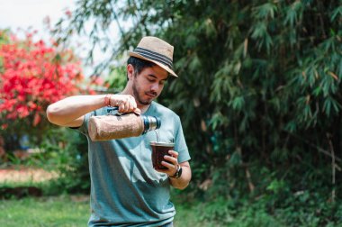 Sakallı, şapkalı ve yeşil gömlekli neşeli bir adam açık hava avlusunda hizmet ediyor. Gülümseyen bir keyif ifadesi. Arjantin ve Uruguay 'ın tipik yerba dostuyla sıcak bir içecek.,
