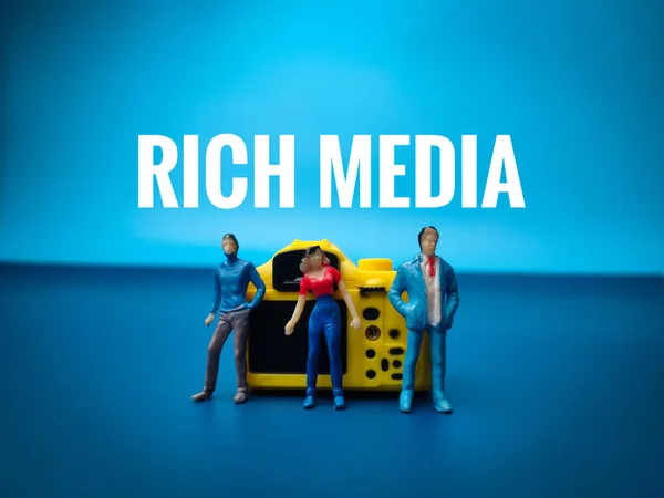 Closeup miniature people and yellow camera with the word RICH MEDIA on blue background.